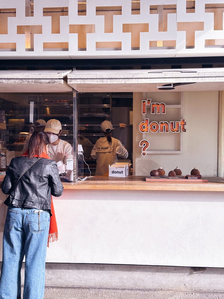 東京甜甜圈店外帶窗口，一名顧客在櫃檯前點餐，店內工作人員正在準備甜甜圈，呈現日常販售場景