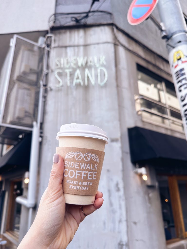 東京街角咖啡店前，一隻手拿著外帶咖啡杯，背景為轉角建築與街景，呈現城市日常片刻